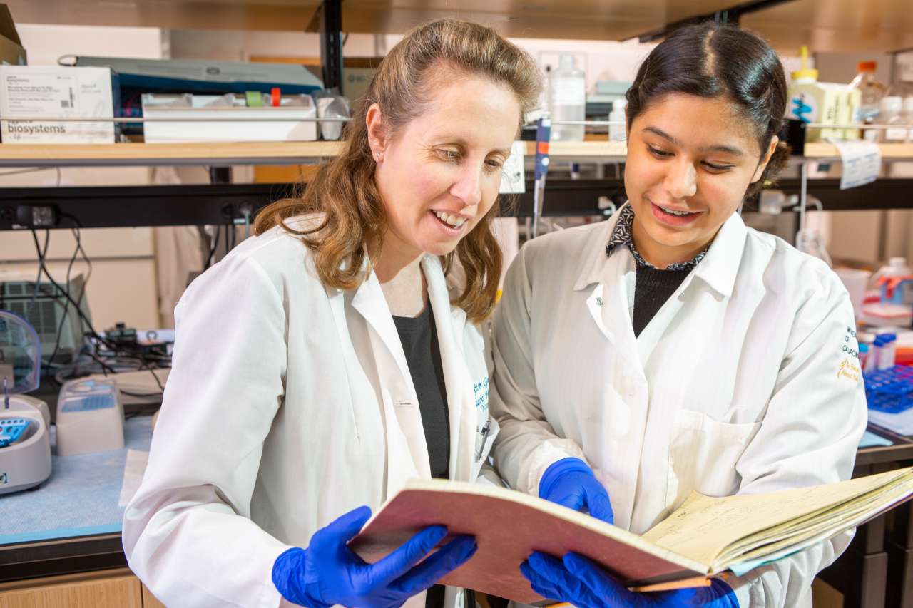  Two smiling women in lab coats and blue gloves examine a book in a lab. The woman on the left has light brown hair, and the woman on the right has dark brown hair.
