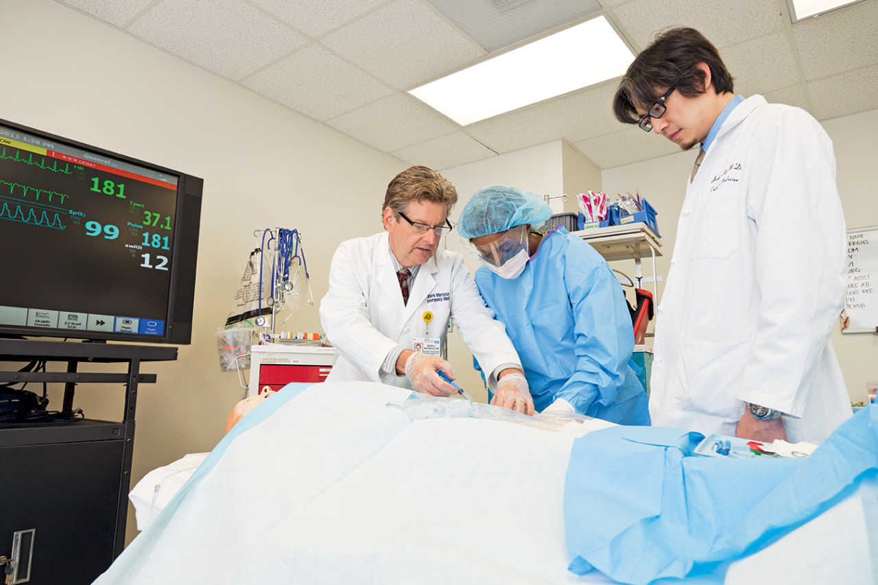  Three medical professionals, one in scrubs, attend to a patient on a bed in a medical setting.