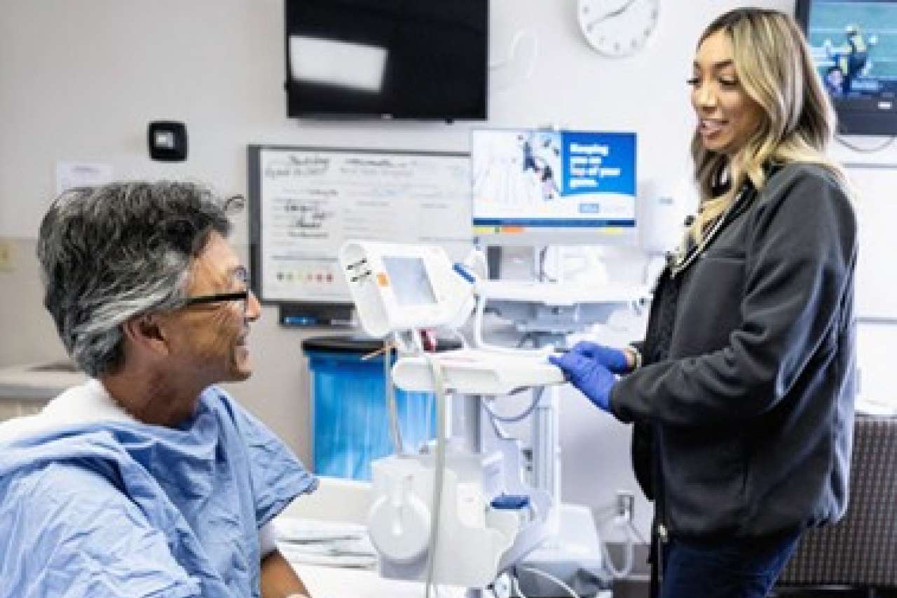 A patient in a hospital gown sits on a bed while a nurse Mariah Gonzalez stands nearby.