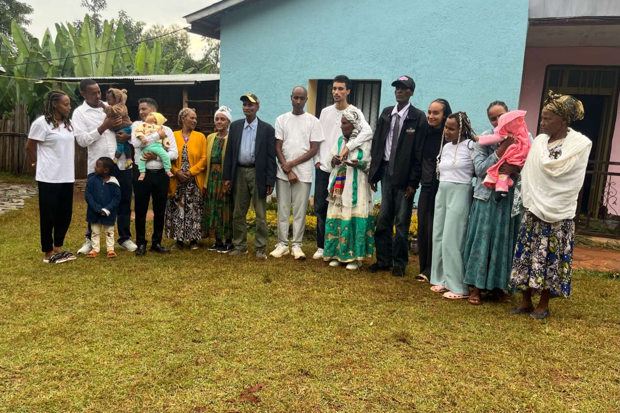 A multigenerational group of about a dozen people, including young children and babies, pose together outdoors in front of a light blue house with lush green vegetation in the background. Several people wear traditional Ethiopian clothing, including colorful embroidered garments and head wraps, while others are dressed in casual or formal Western attire.