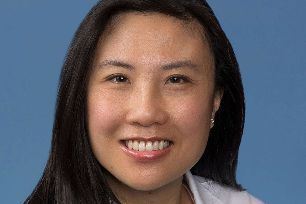 Kara Chew smiling in a white coat against a blue background.