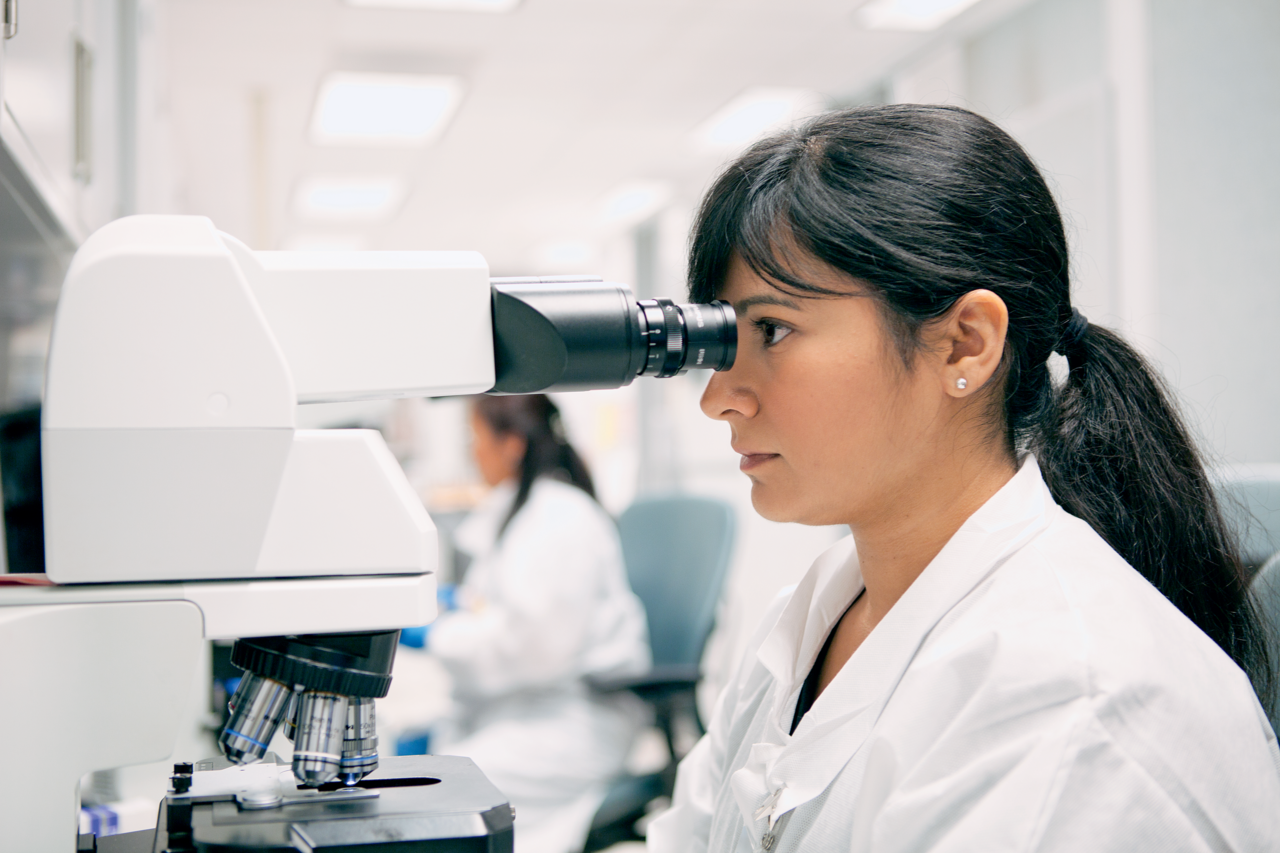 A technician wearing white lab coat looking into a microscope in a laboratory, with another person in the background.
