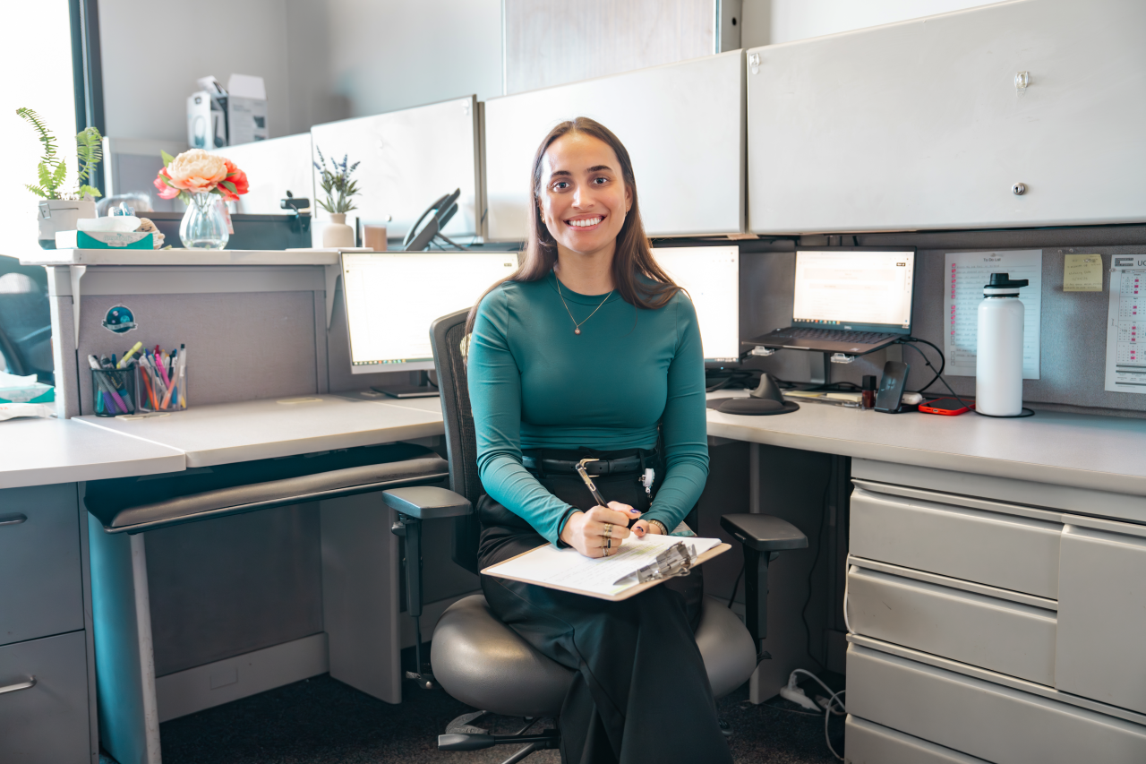 VA Administrative Staff seated at her desk in an office environment. She is smiling and holding a clipboard.