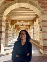 A woman with dark brown curly hair and glasses smiling, standing underneath the sunlit arches