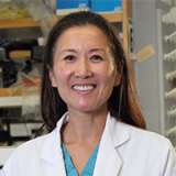 A smiling headshot of Dr. Linda Liau, an Asian woman with long dark hair, wearing a white lab coat over light blue scrubs, in a laboratory setting.