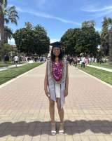 Graduation photo of Camille, who is wearing a black cap, purple flower lei, white dress, stole, and shoes.