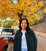 Diana, who has brown hair and is wearing a white shirt and black jacket, smiling in front of a tree with yellow leaves.