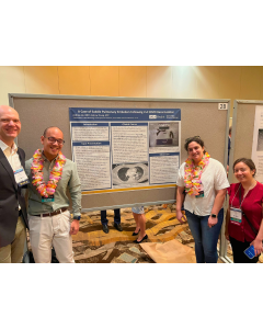 Four people wearing leis stand next to a research poster at a conference.