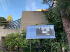 Blue historical plaque with black-and-white photo outside concrete building and trees.