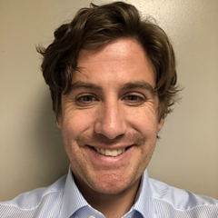 A headshot of Charles McTavish who has light brown hair and green eyes, smiling and wearing a light blue striped shirt.