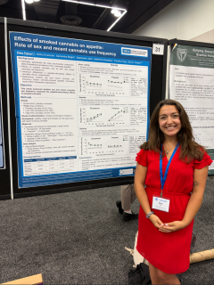 Photo of Elisa Pabon, PhD who has brown hair and is wearing a red dress, standing in front of a blue and white research poster.