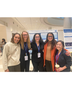 Five women pose together at a conference, smiling in front of a research poster.
