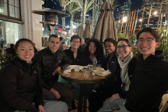 A group of six friends gathered around a table at night, smiling and enjoying food.