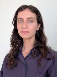 Woman with dark wavy hair and blue eyes, neutral expression on white background.