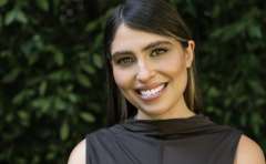 Smiling woman with dark hair wearing a black top against leafy green background