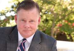 Man in a suit with a tie, sitting outdoors amidst greenery.