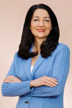 Johnese Spisso, a woman with dark hair, smiles with her arms crossed. She wears a blue textured sweater and a pearl necklace.