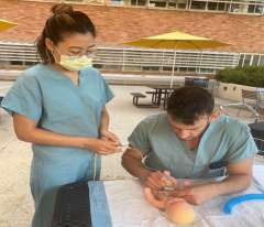 Two healthcare workers in scrubs practicing suturing on a model at an outdoor table.