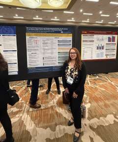 Dr. Victoria Rodriguez, who has brown hair and glasses, standing in front of research poster.