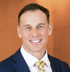 Patrick Loney in a suit with a striped tie, against a soft orange background.