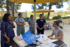 Person seated at picnic table shows papers while four others stand listening under umbrella