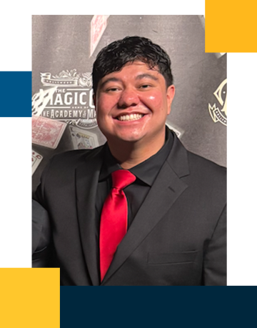 Josh is smiling while standing in front of a magic castle background in a black suit with a red tie