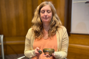 Diana Winston, with closed eyes, meditates holding a singing bowl and mallet. She has long, wavy blonde hair and wears an orange top and a light cardigan.