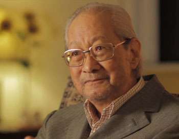 Elderly man sitting in a cozy setting, wearing glasses and a suit.