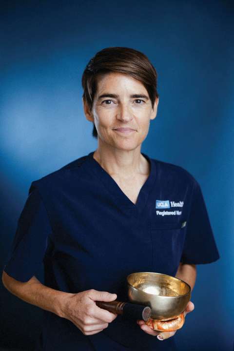 Lindsay Brant holding a singing bowl, wearing a dark medical outfit, against a blue background.