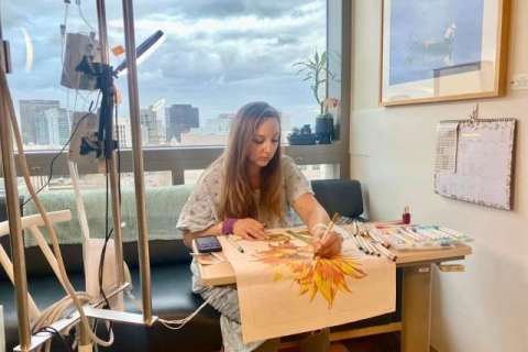 Person painting a sunflower canvas at a table by a window with city skyline