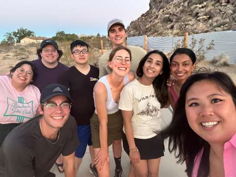 Nine people smiling in a group selfie outdoors with rocky hills behind