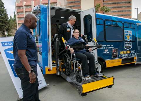 A person in a wheelchair is being assisted onto the Operation Mend bus by a bus driver, while another person stands by. 