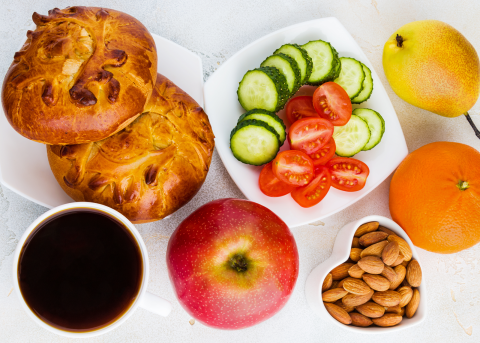 Balanced meal with bread rolls, sliced cucumber and tomato, almonds, fruit, and coffee, promoting healthy nutrition habits.