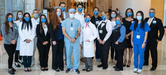 A group of healthcare professionals and staff stand together in a hospital lobby, wearing a mix of scrubs, lab coats, and business attire.