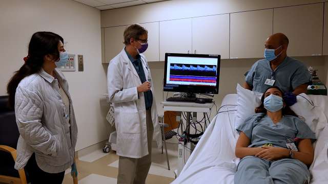 Hospital team of masked clinicians reviewing heart monitor beside a patient in bed.