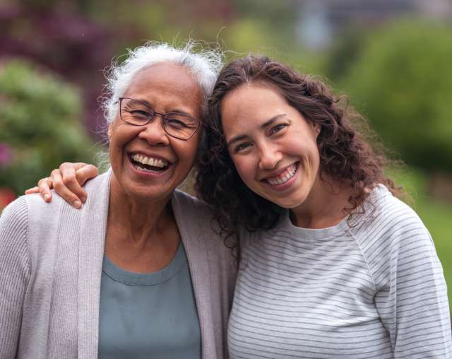 A candid, medium close-up shot shows two women of different generations embracing and smiling at the camera outdoors.
