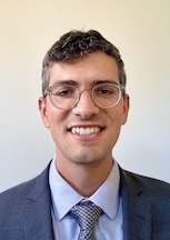  A headshot of Daniel Stokes, a smiling man with short curly hair and glasses, wearing a suit and patterned tie.