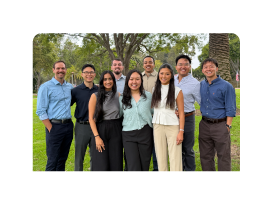Nine adults standing and smiling outdoors on grass beneath trees.