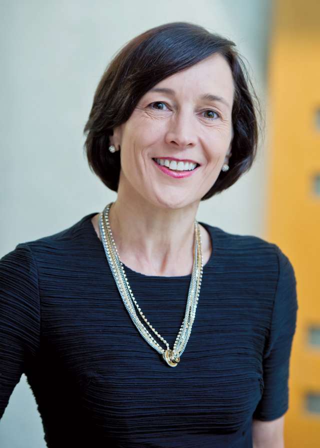 Woman wearing a black dress and layered necklaces, standing in a modern setting.