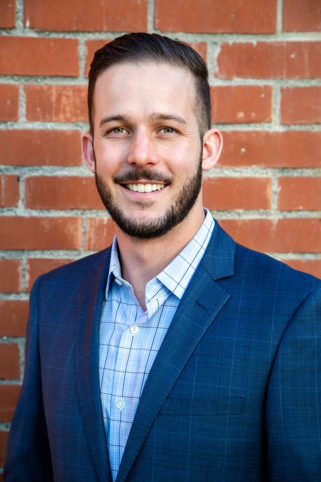 Matt Flesock in a blue suit and checkered shirt stands in front of a brick wall.