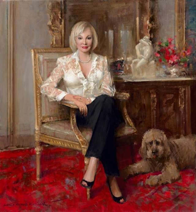 Iris Cantor seated in a lace blouse and black pants poses with a dog on a red carpet.