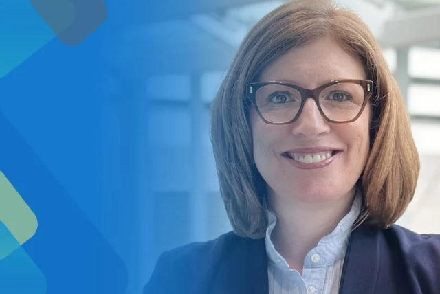 Headshot of Marylyn Ritchie, she's wearing brown glasses, a light blue button up shirt, and a dark blue suit coat.