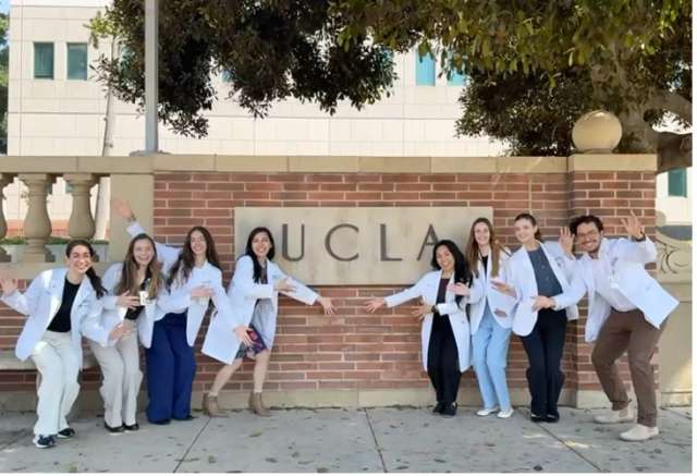 April Armstrong Lab fellows wearing their lab coats posing in front of a UCLA wall engraving in campus