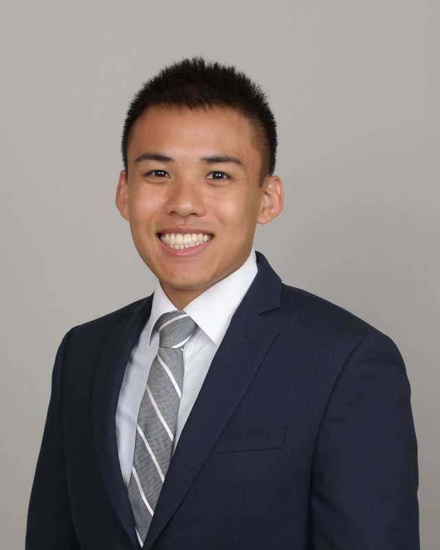  A headshot of Kevin La, a smiling man with short dark hair, wearing a dark suit and a striped gray and white tie.