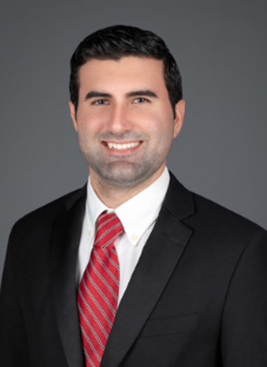 Professional headshot of a person with short dark hair, wearing a black suit jacket, white shirt, and red striped tie, facing forward against a gray studio background.