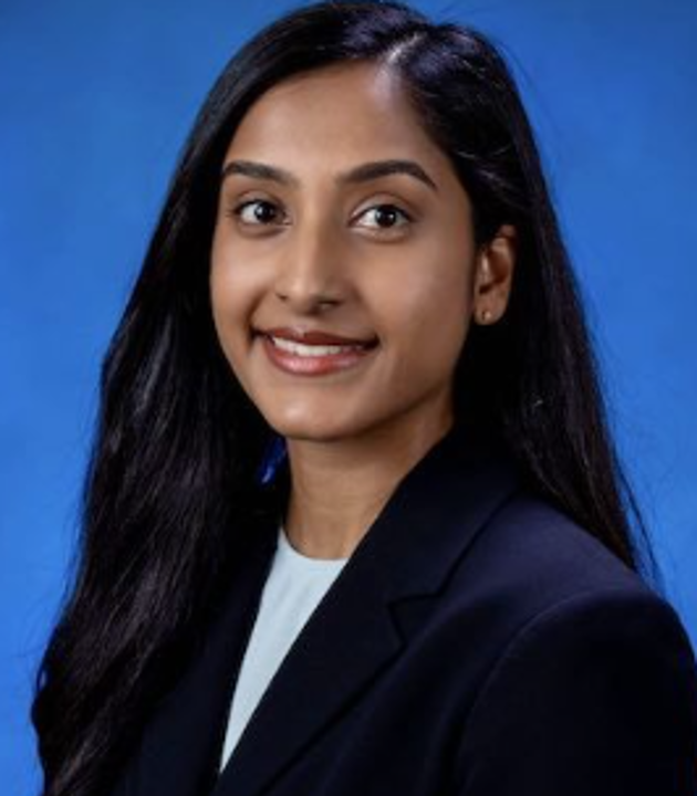 Professional headshot of a person with long straight hair, wearing a dark blazer over a light-colored top, facing forward against a solid blue background.