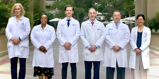 A group photo of six thoracic surgery team members wearing white lab coats, standing outdoors on a pathway with trees and umbrellas in the background. 