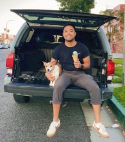 Man sitting on open SUV tailgate holding an ice cream cone with a corgi beside him.
