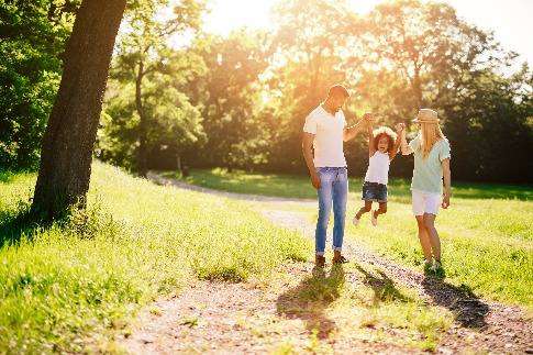 a father, child, and mother holding hands in an outdoor setting. there are grass, trees, and sunshine.