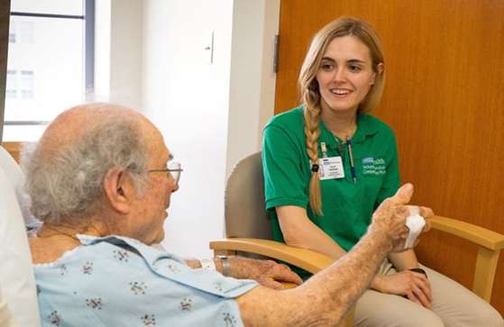 A young woman speaking with an older patient resting in his bed